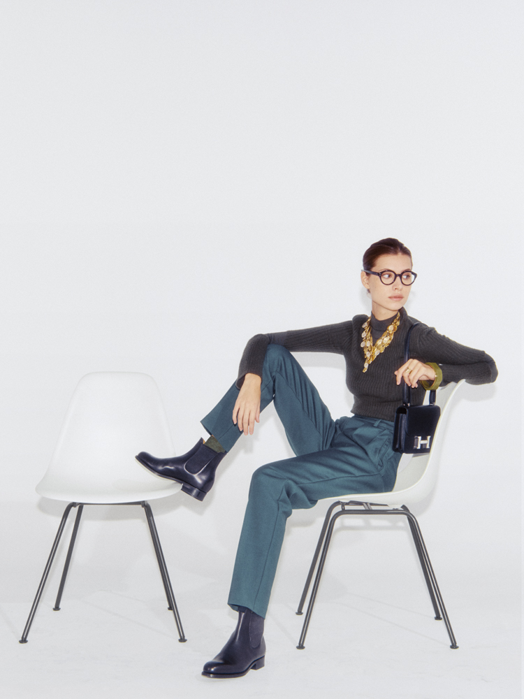 Woman in teal trousers and dark sweater sits on white chair, wearing round glasses and holding small black handbag