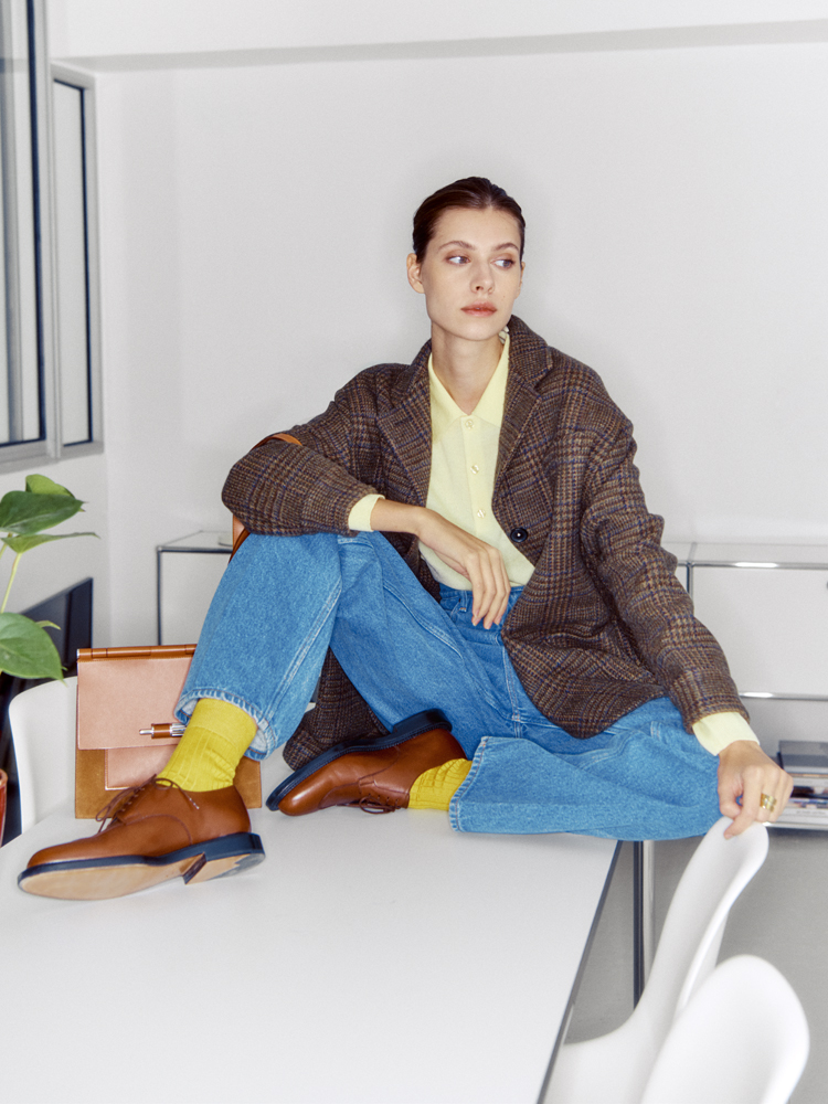 Woman in oversized brown plaid blazer, yellow polo, blue jeans and brown shoes sits casually on white desk