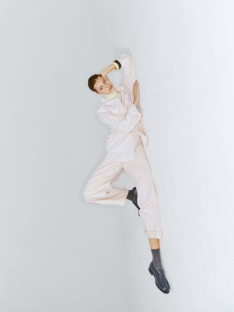 Person in pale pink oversized suit and turtleneck jumping mid-air against white background, wearing dark boots and bangles
