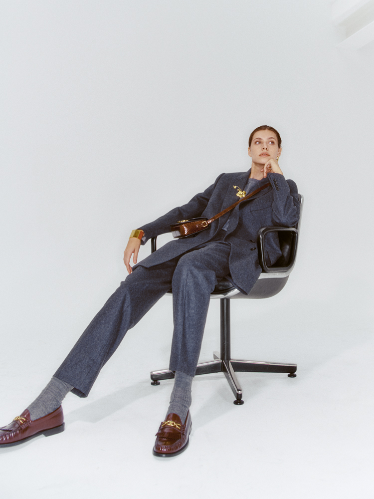 Model in navy suit sits in black chair wearing brown leather accessories and chain-link gold jewelry