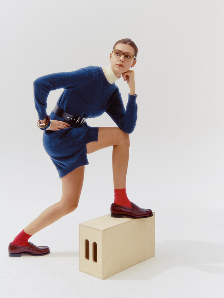 Model in navy belted dress with white turtleneck, red socks, burgundy loafers and glasses poses with foot on wooden block