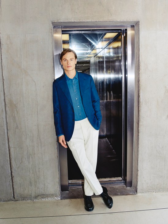 Model wearing blue blazer, denim shirt, white trousers and black shoes standing in elevator