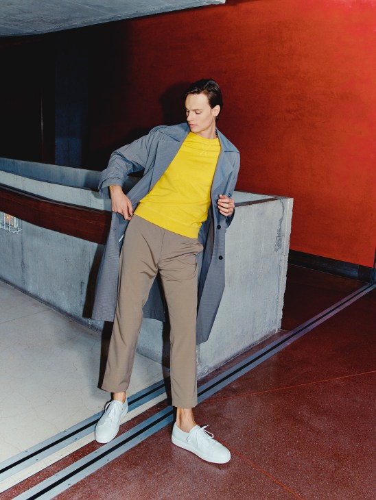 Model wearing blue coat, yellow sweatshirt, beige trousers and white trainers against red and grey architectural backdrop.