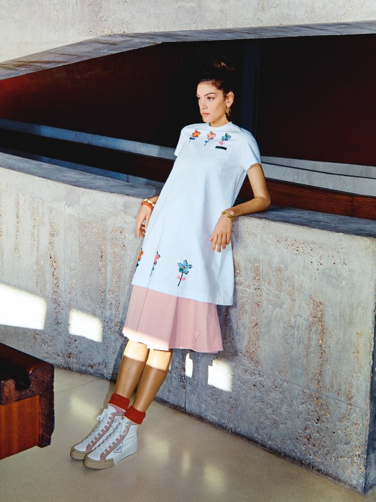 Model wearing Prada t-shirt dress, skirt, trainers and socks with Patou earrings and Saint Laurent bracelets.