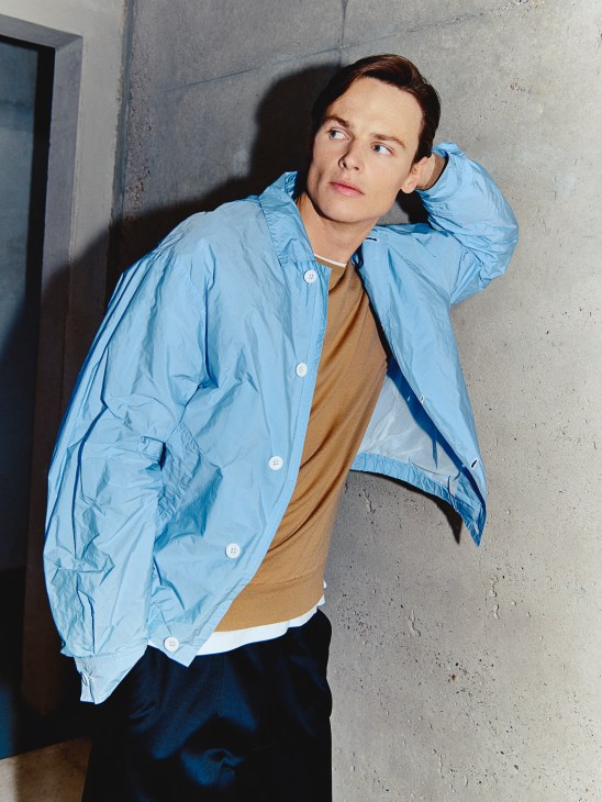 Male model wearing a light blue Scye jacket over a mustard John Smedley jumper and white t-shirt, posing against a concrete wall.