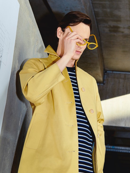 Model wearing a yellow Mackintosh coat and striped Norse Projects jumper with yellow Persol glasses.