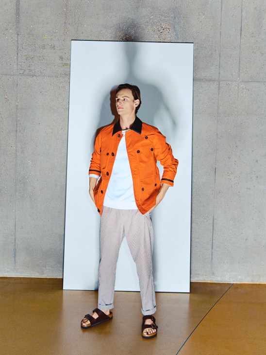 Man in orange Drake's jacket, Colmar sweatshirt, A Kind of Guise trousers and Birkenstock sandals against white backdrop.