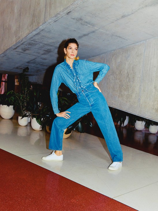 Model wearing blue denim jacket and trousers with white sneakers, posing in an industrial venue.
