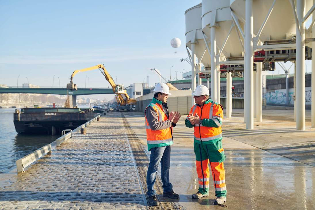 Two construction workers in high-visibility vests and hard hats at a waterfront construction site for the Grand Paris Express metro project.
