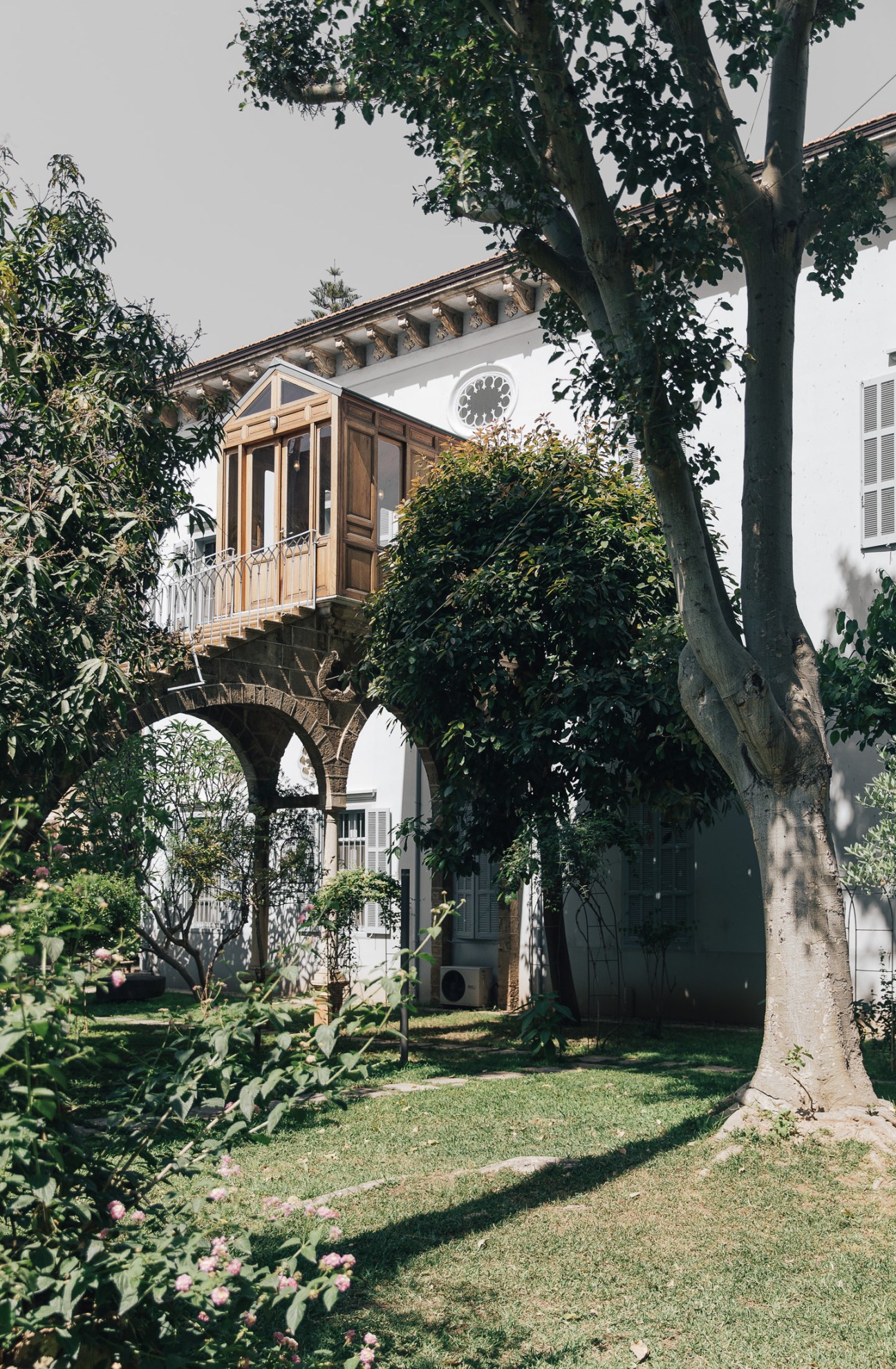 A 19th-century mansion with ivy-covered walls, arched entryway, and wooden balcony overlooking a lush garden with flowering plants.