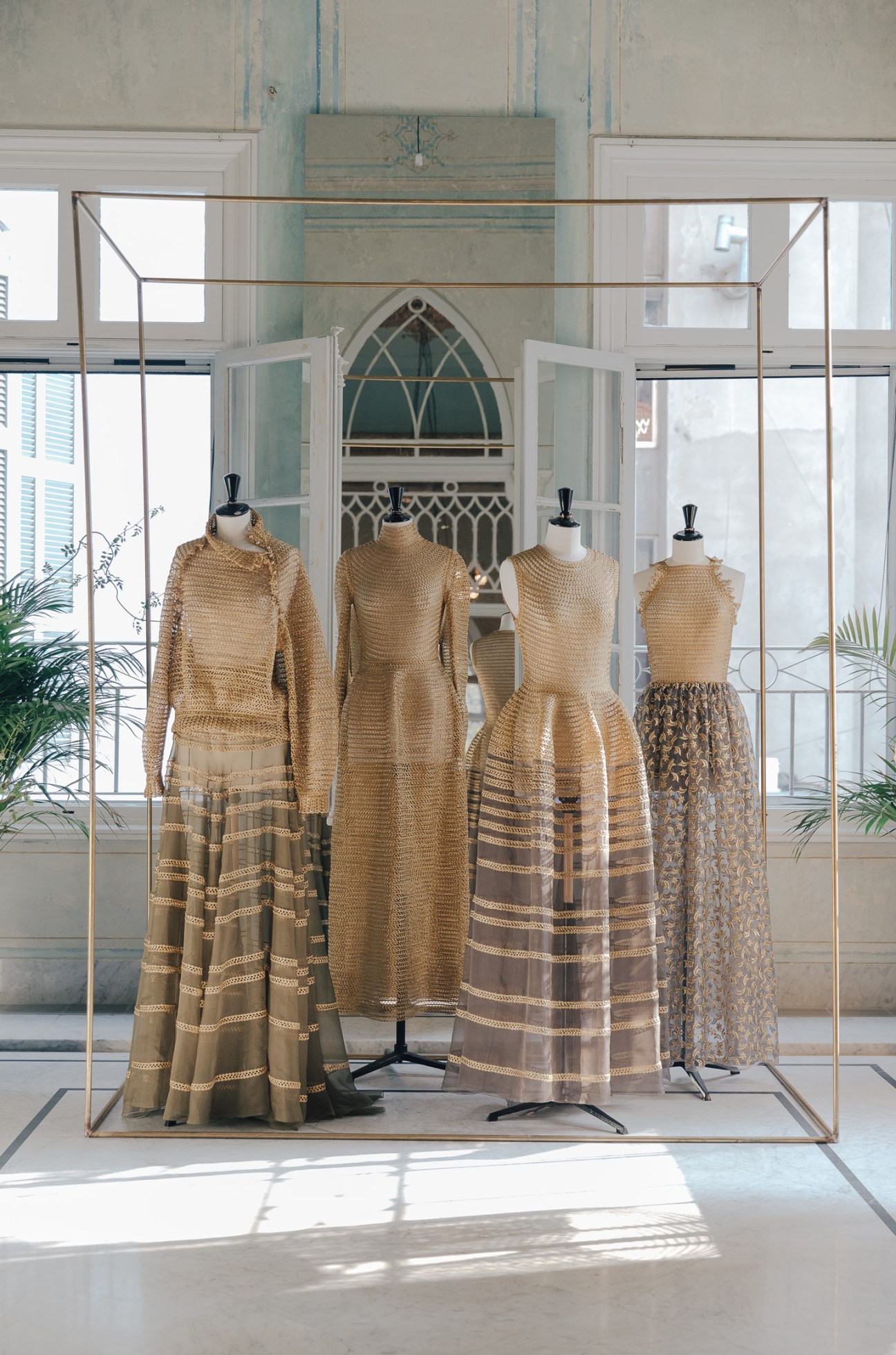 Four mannequins displaying Rabih Kayrouz gowns in gold and neutral tones with decorative details in a bright salon with arched windows.