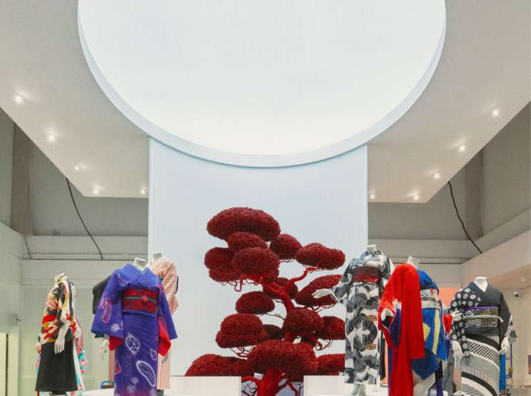 Exhibition display with colorful kimono garments on white platforms around a central red sculptural tree