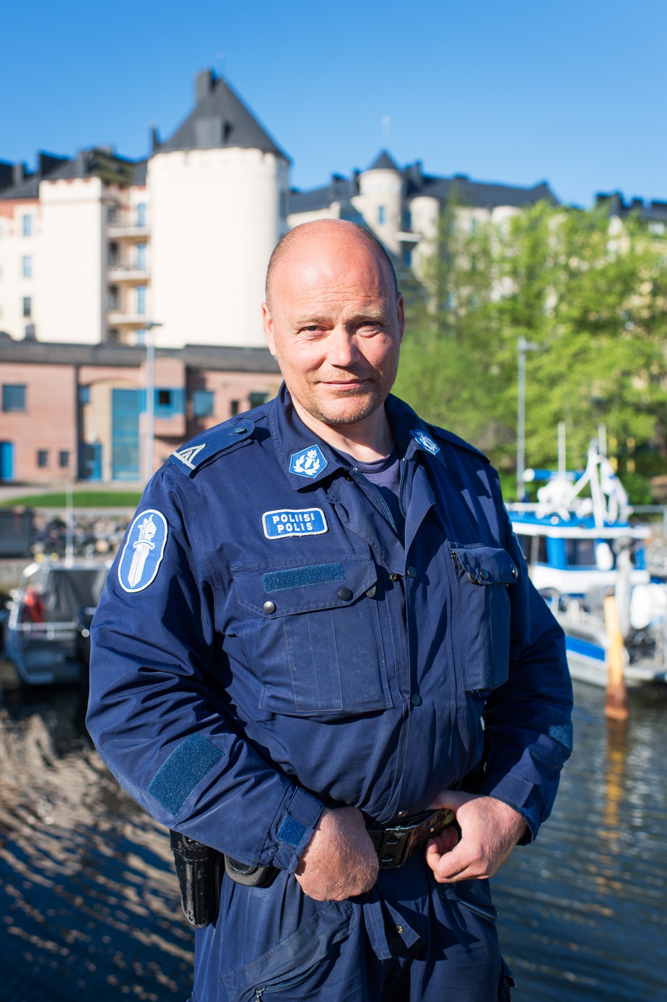 Sergeant Jarmo Niininen of Helsinki water police stands in uniform at the waterfront with boats and historic architecture behind him.