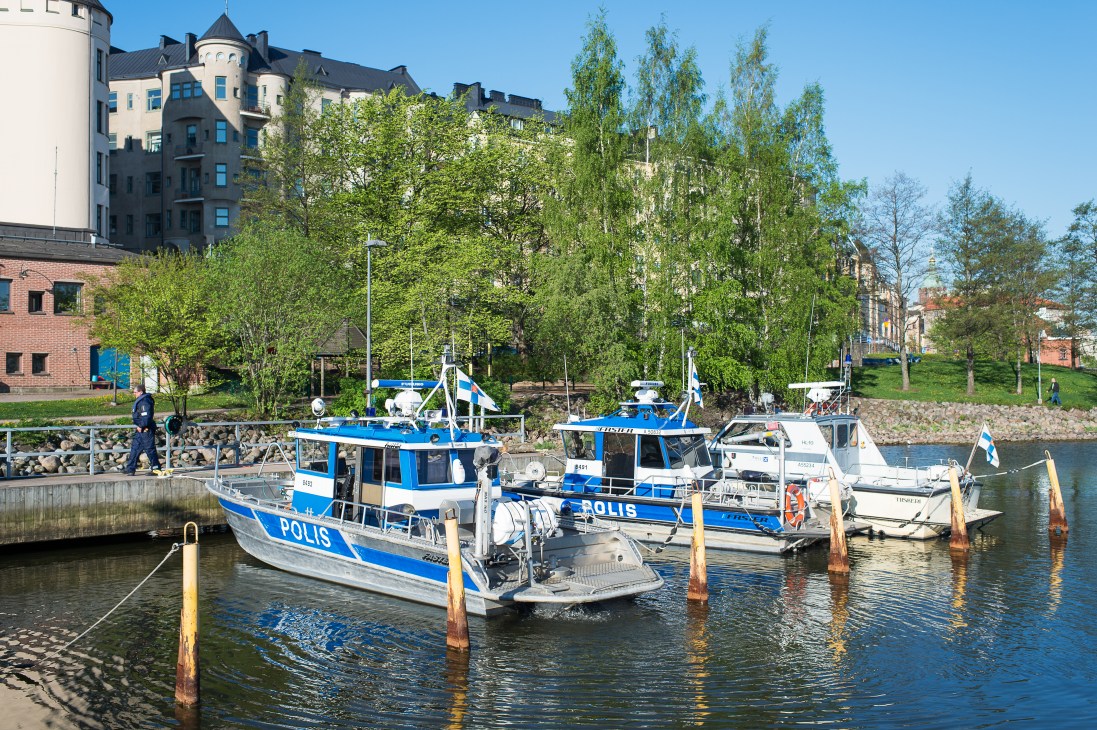 Blue and white police boats moored at a dock in Helsinki with historic waterfront buildings in the background.