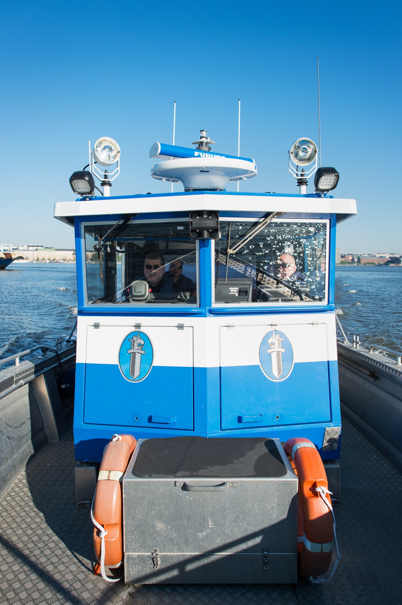 Helsinki water police boat with officers visible through windows, moored at a dock with water and cityscape in background.
