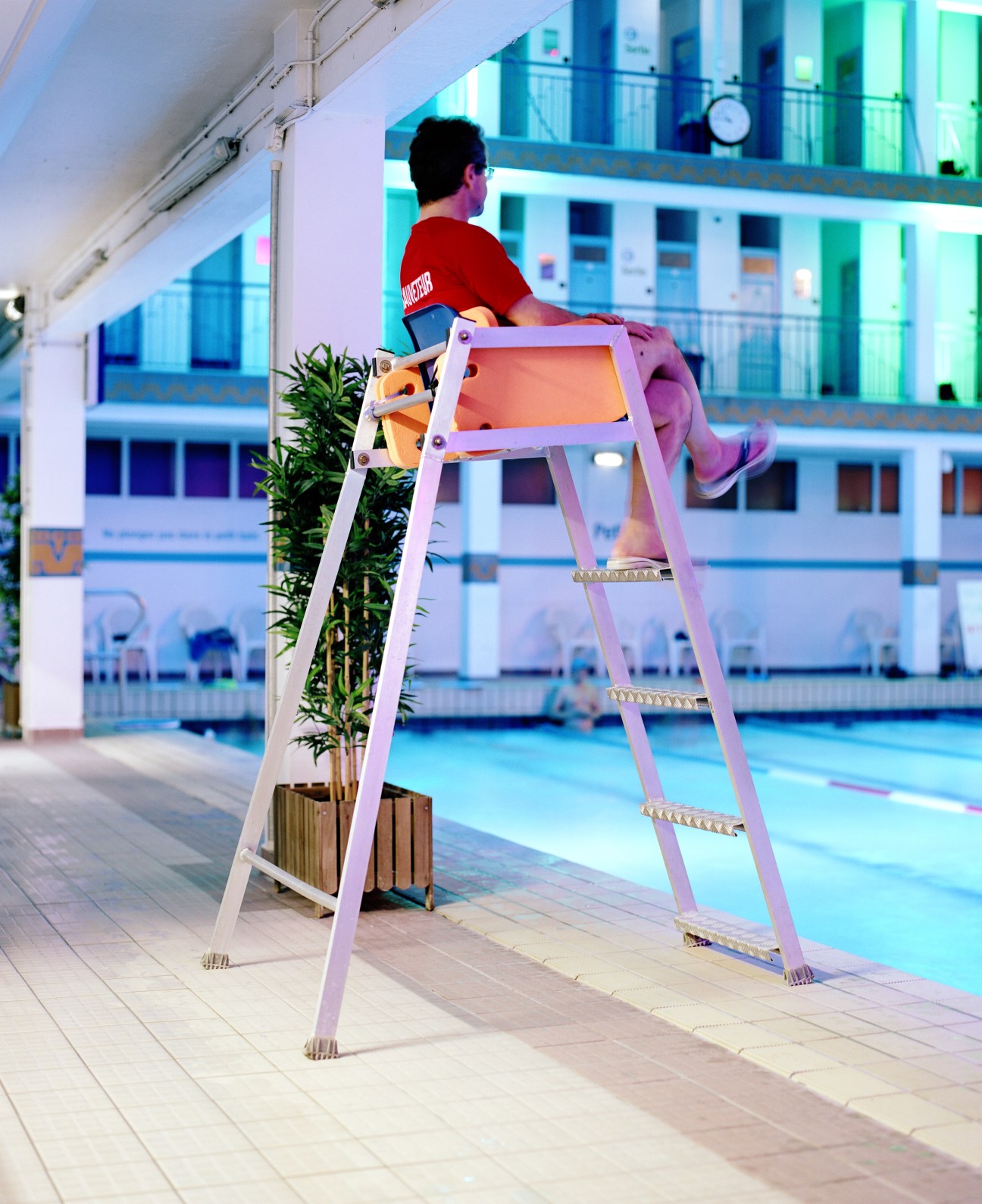Lifeguard standing on elevated chair overlooking an indoor swimming pool with neon-lit water.