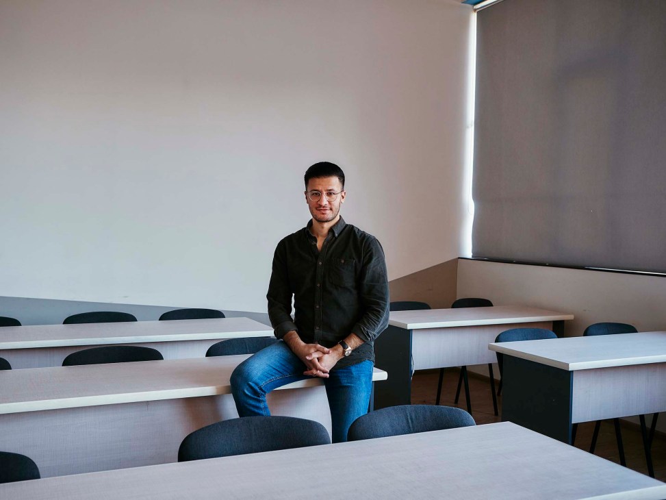 Narek Aramjan sits on a desk in a modern office space with multiple white tables and blue chairs.