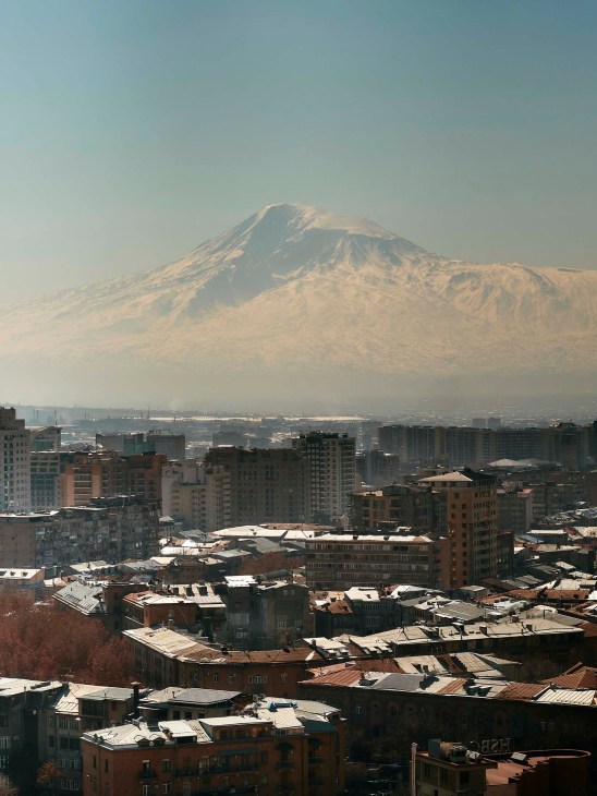 Snow-capped Mount Ararat rising above residential buildings in Yerevan, Armenia, with hazy atmospheric conditions