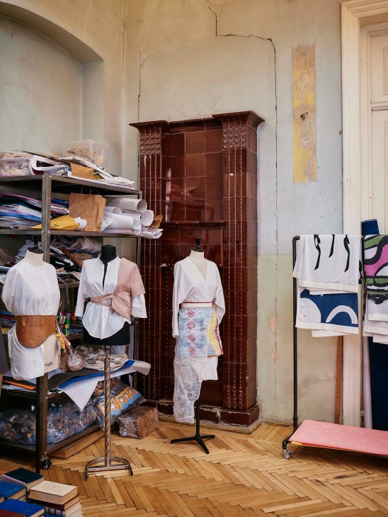 Clothing displayed on mannequins and racks inside Tumo Studios creative workshop with ornate wooden furniture and herringbone flooring.