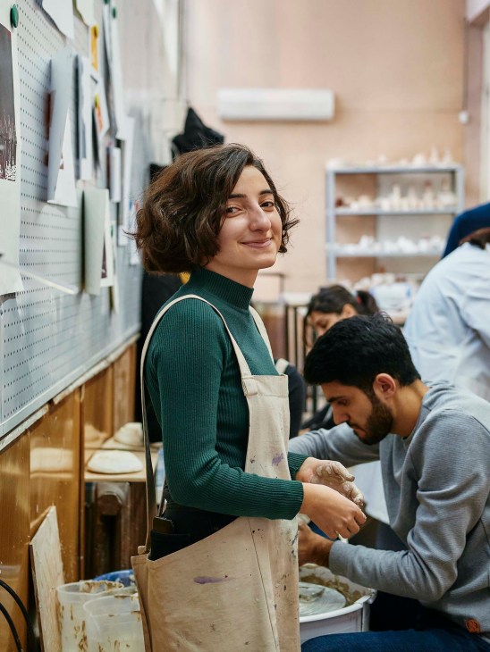 Young woman in green sweater and apron smiles at clay station during pottery class at Tumo Studios in Yerevan.