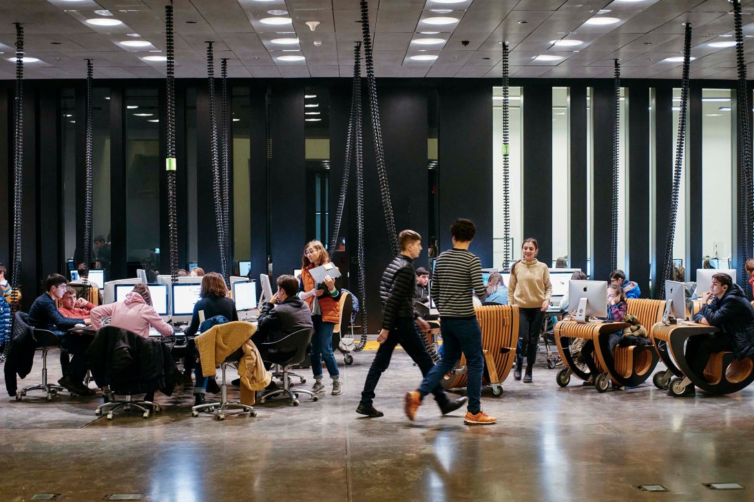 Students and visitors move through a modern creative technology center with computers, hanging sculptural elements, and wooden seating at Tumo.