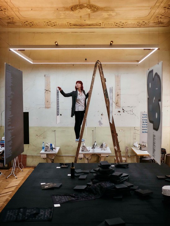A woman stands on a wooden ladder arranging graphic design work in a modern studio space with neon lighting.