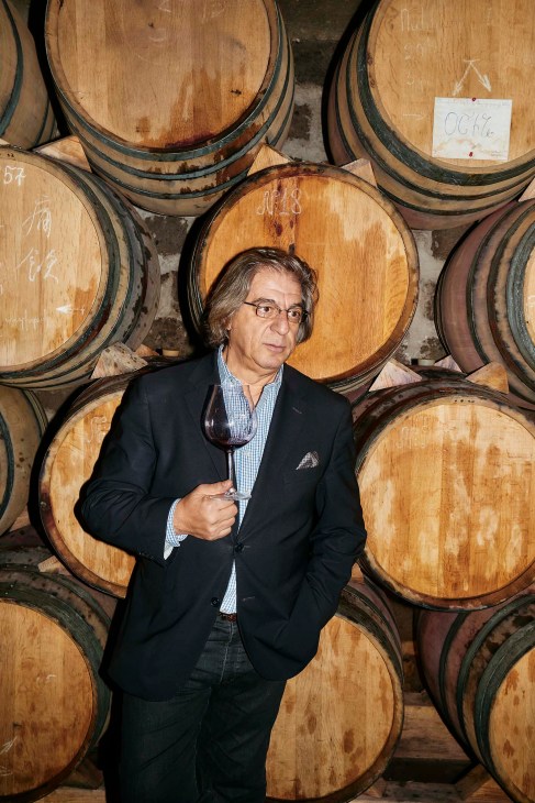 Vahe Keushguerian, winemaker, holds a glass of wine while standing among wooden casks in a winery.