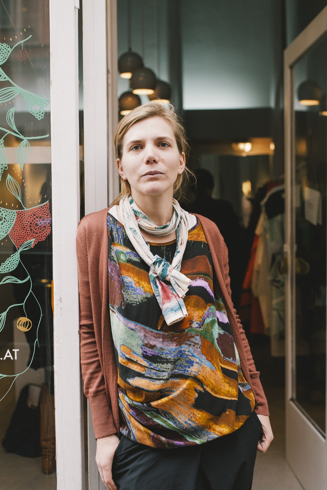 Woman standing in front of a storefront wearing a rust-colored cardigan and patterned top.