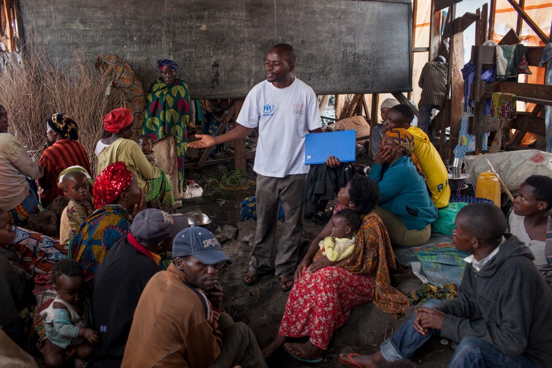 Humanitarian worker in UNHCR shirt holds laptop while speaking with internally displaced people gathered in a shelter in DRC refugee camp.