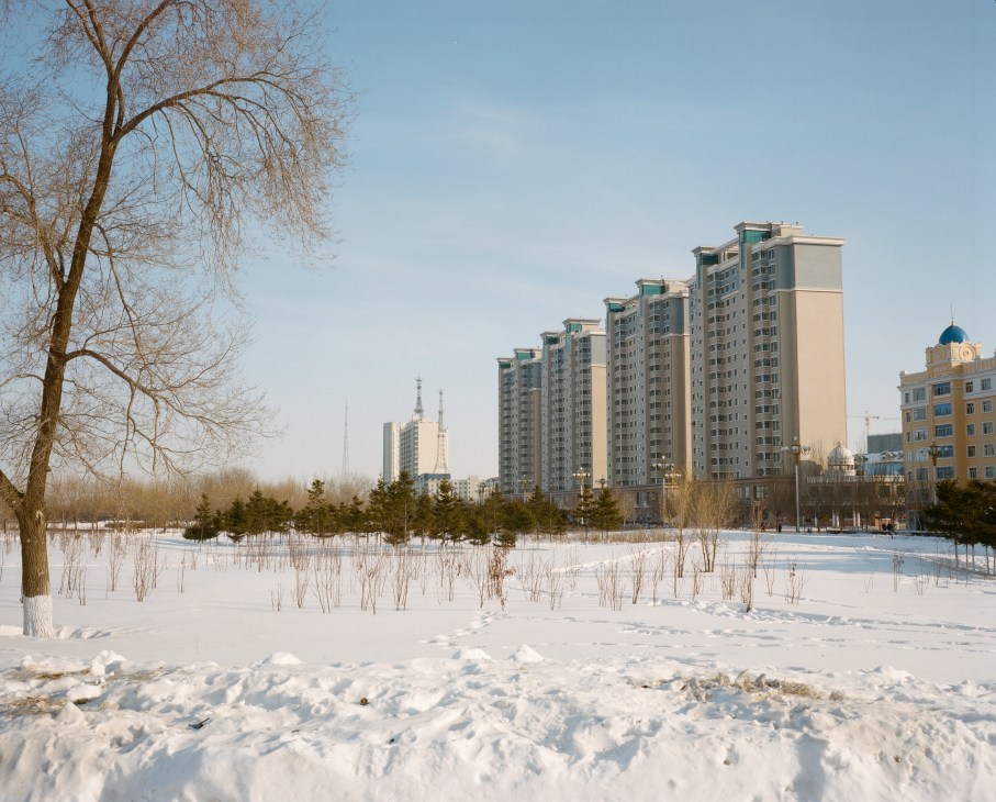 Snow-covered Amur River with frozen landscape and bare trees facing modern apartment buildings and churches of Heihe, China.
