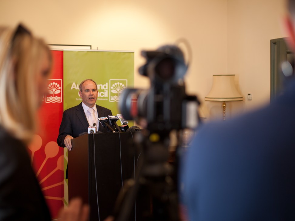 Man in dark suit speaking at podium with microphones during press conference.