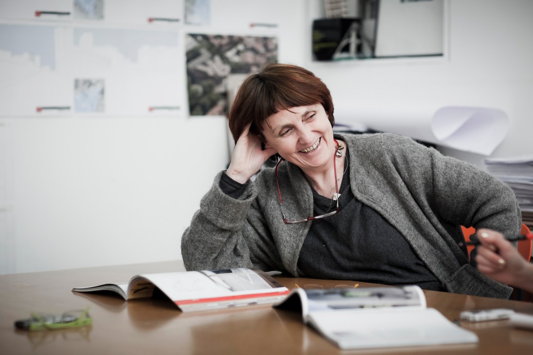 Shelley McNamara, co-founder of Grafton Architects, smiles at her desk in an office with design sketches on the wall behind her.