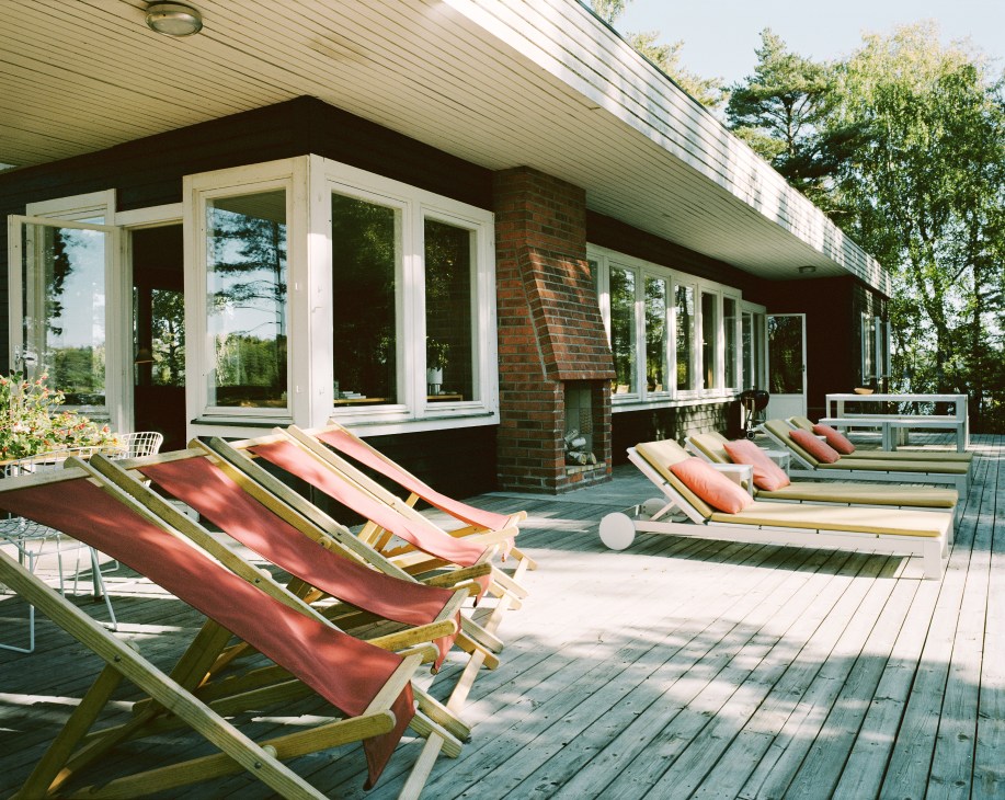 Wooden deck with red cushioned loungers and large windows of a brick house surrounded by trees.