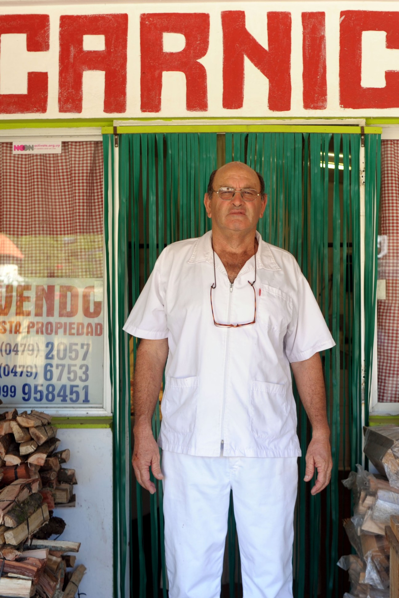 Juan Izaguirre, village butcher, standing in front of his shop with red signage and green corrugated doors.