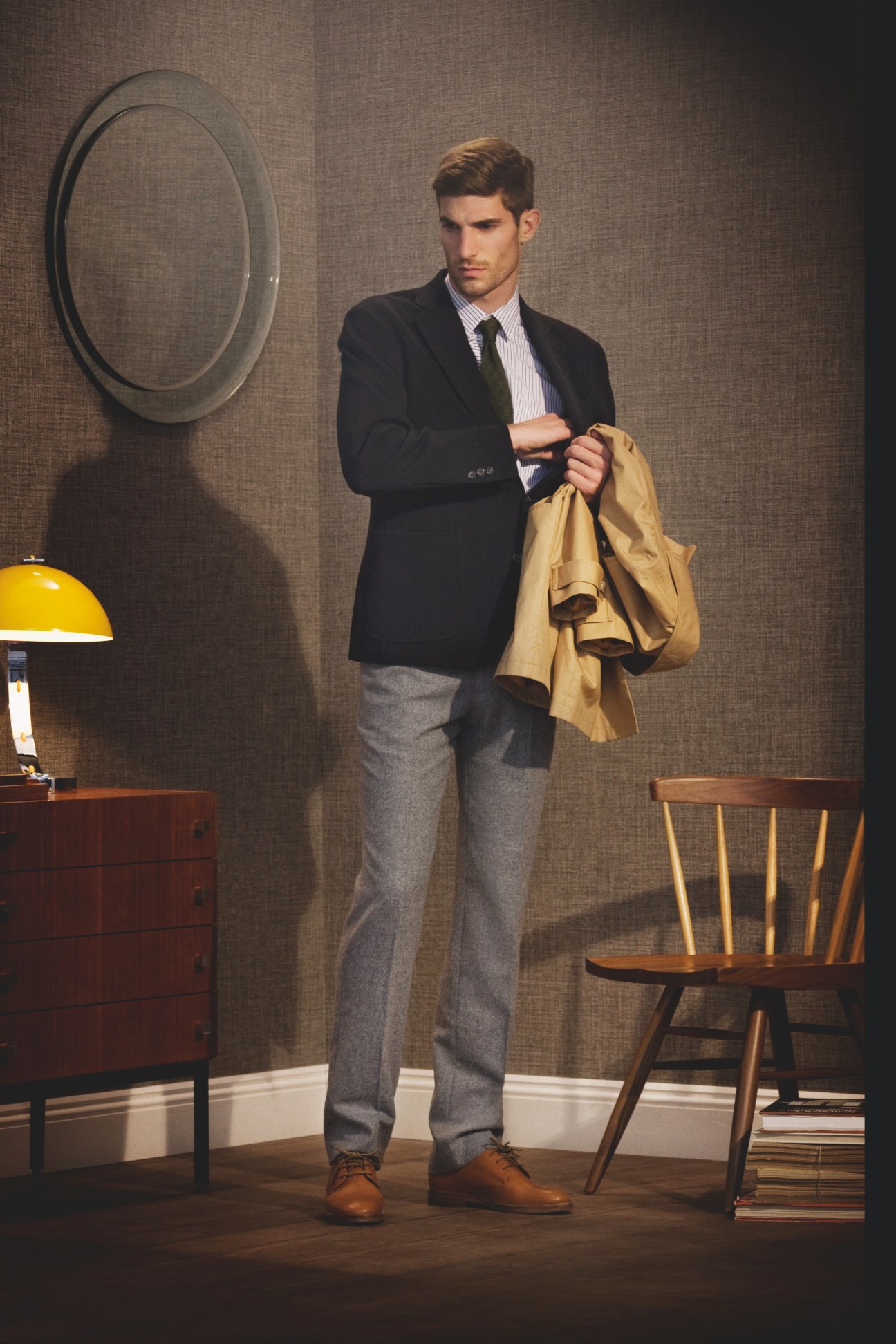 Man in navy blazer, striped shirt and gray trousers holding tan coat, standing in mid-century modern interior.