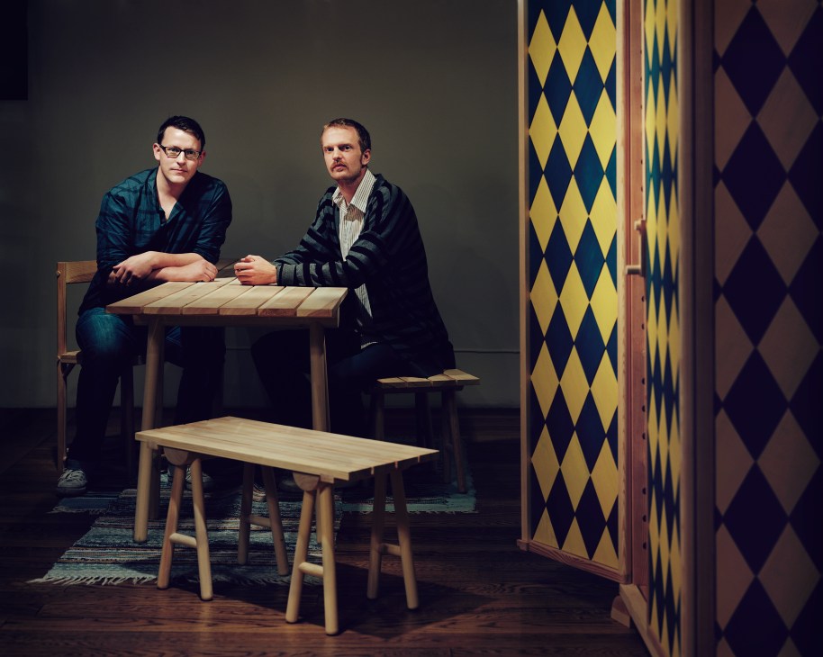 Two men pose with wooden furniture pieces in a studio setting with geometric patterned panels.