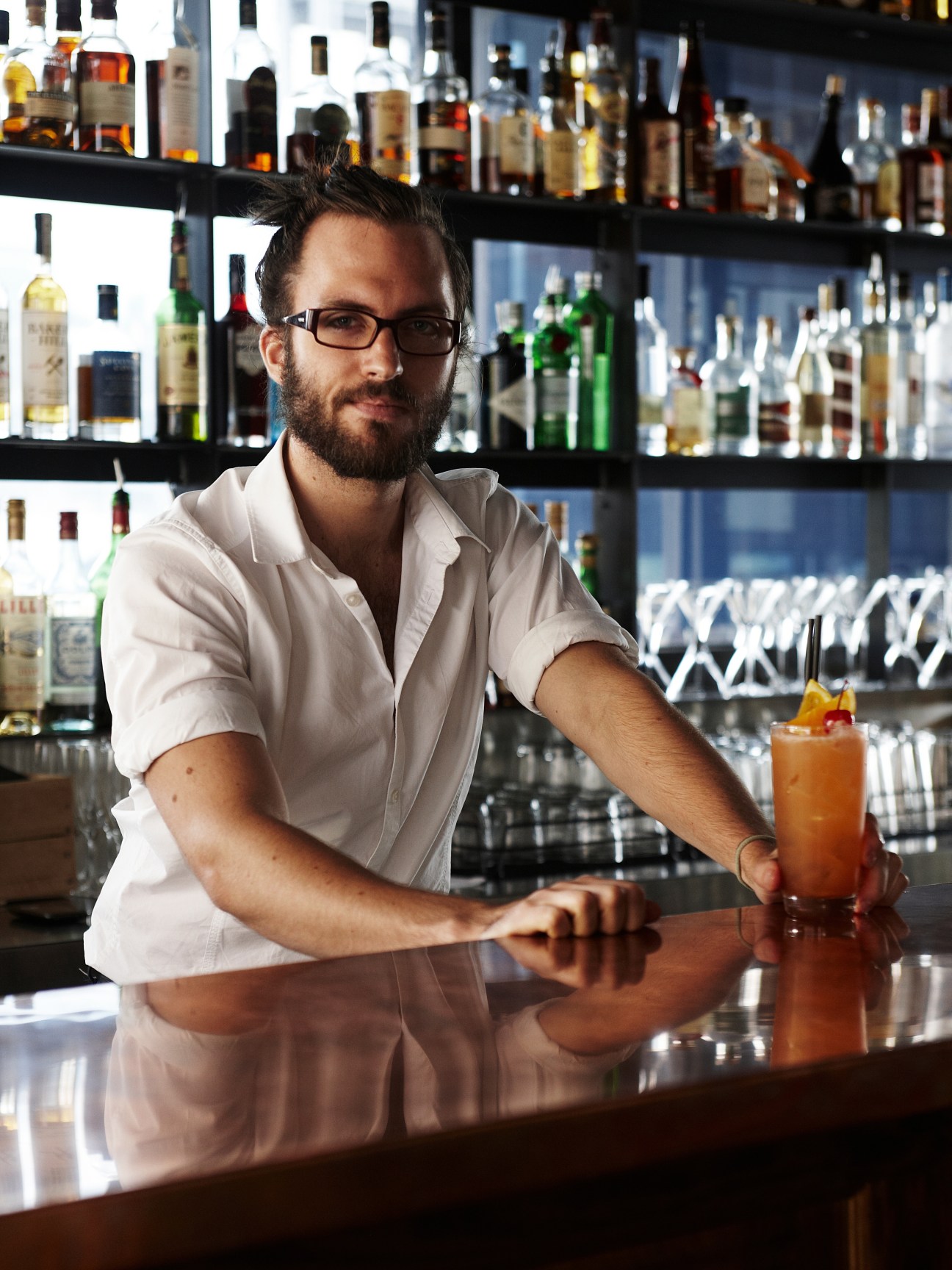 Bartender in white shirt pouring a cocktail behind a bar lined with bottles.