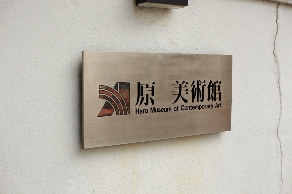 Metal plaque with Hara Museum of Contemporary Art logo and bilingual signage in English and Japanese characters on white wall.
