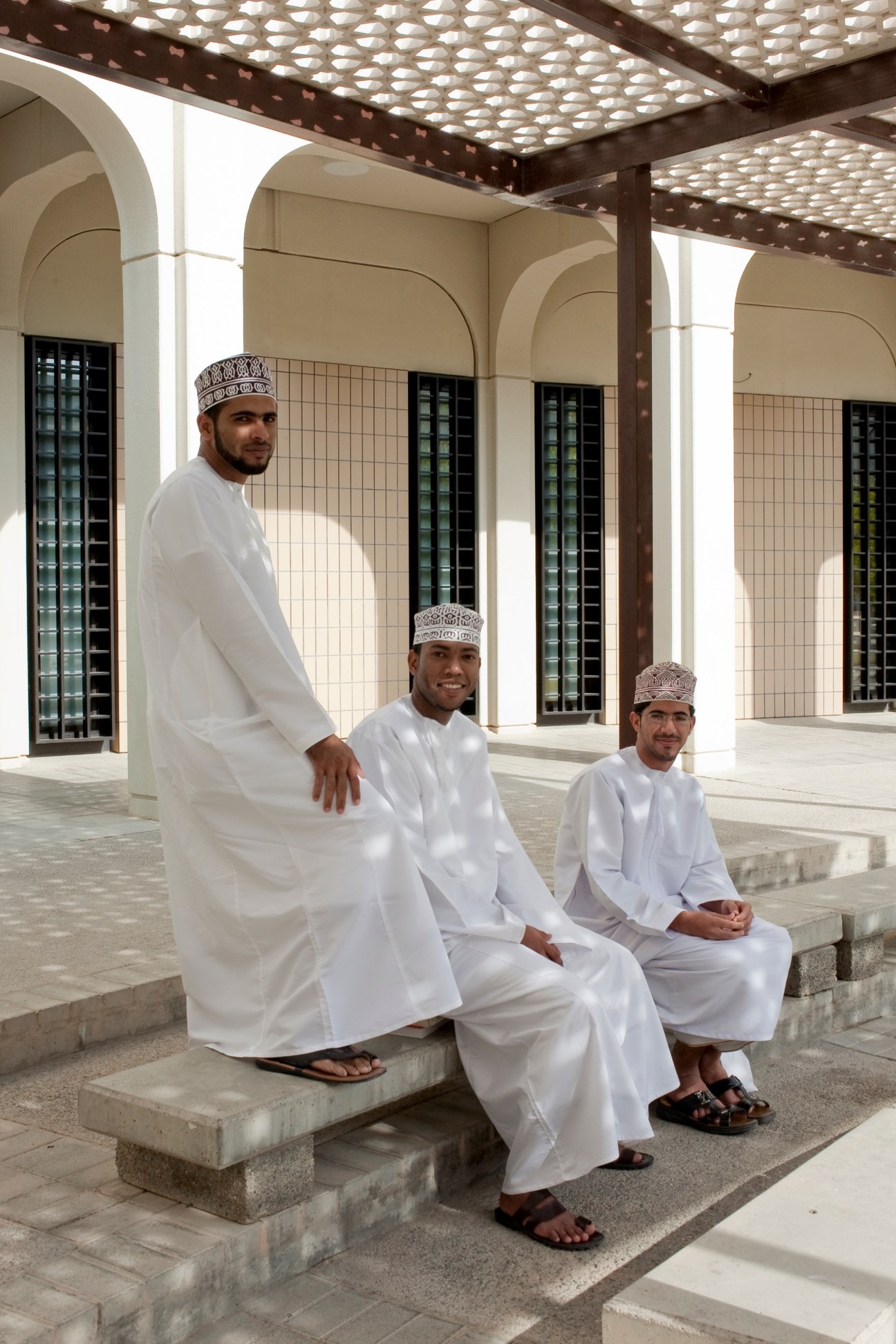 Three male students in white traditional Omani dress pose in a mosque courtyard with white arches and patterned wooden ceiling.
