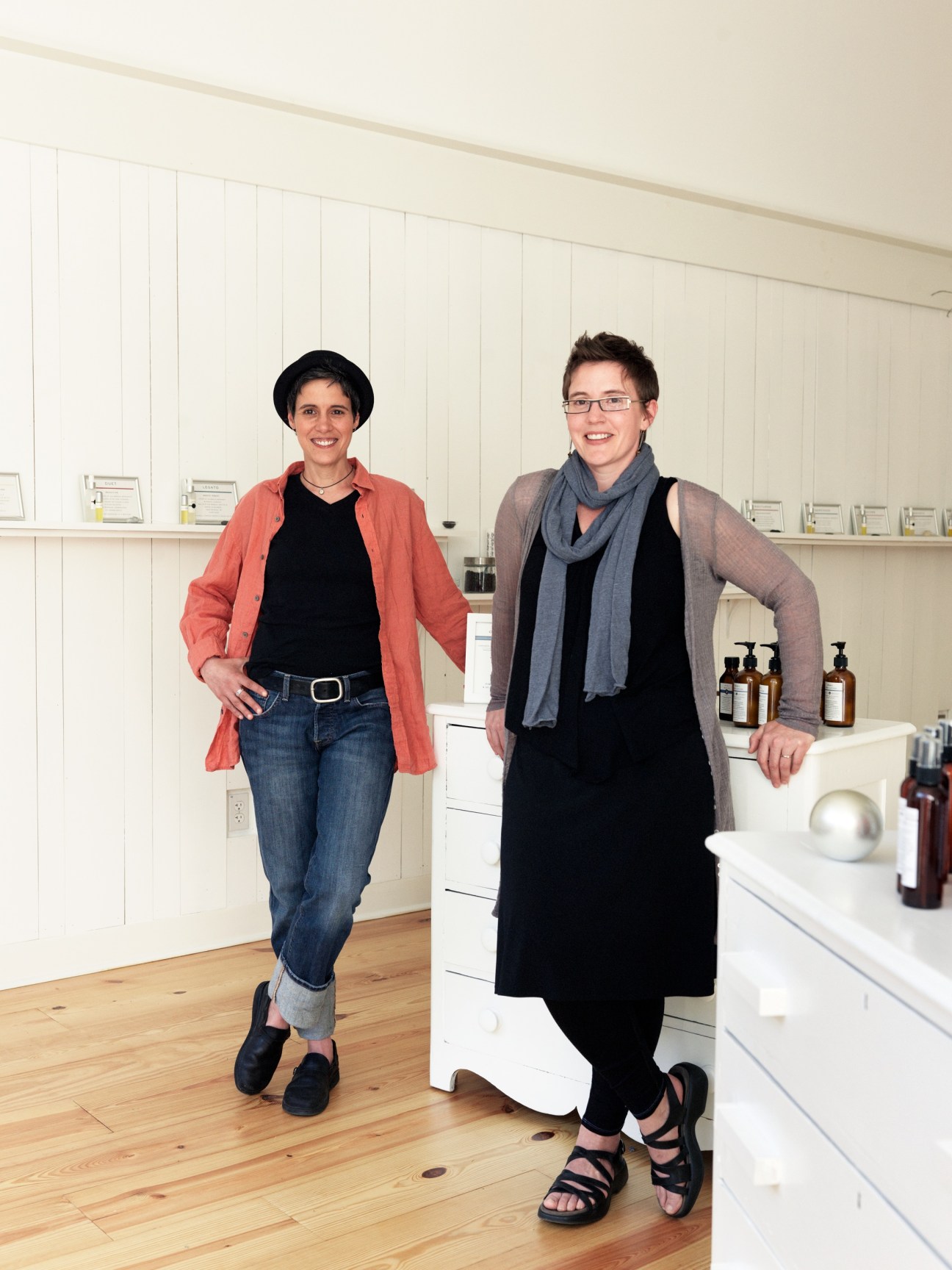 Carolyn Mix and Darcy Doniger stand in their bright perfumery studio with white shelving and amber bottles.