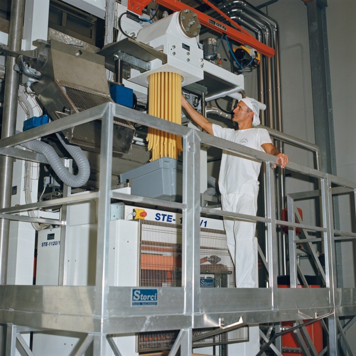 Worker guiding golden penne pasta through a bronze die machine at a pasta factory.