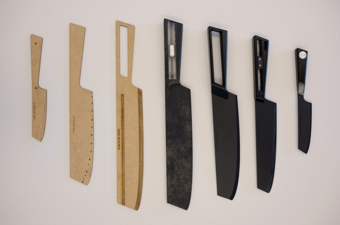 Seven knife prototypes in varying designs, displayed against a white wall, showing progression from natural cork and wood to black finishes.