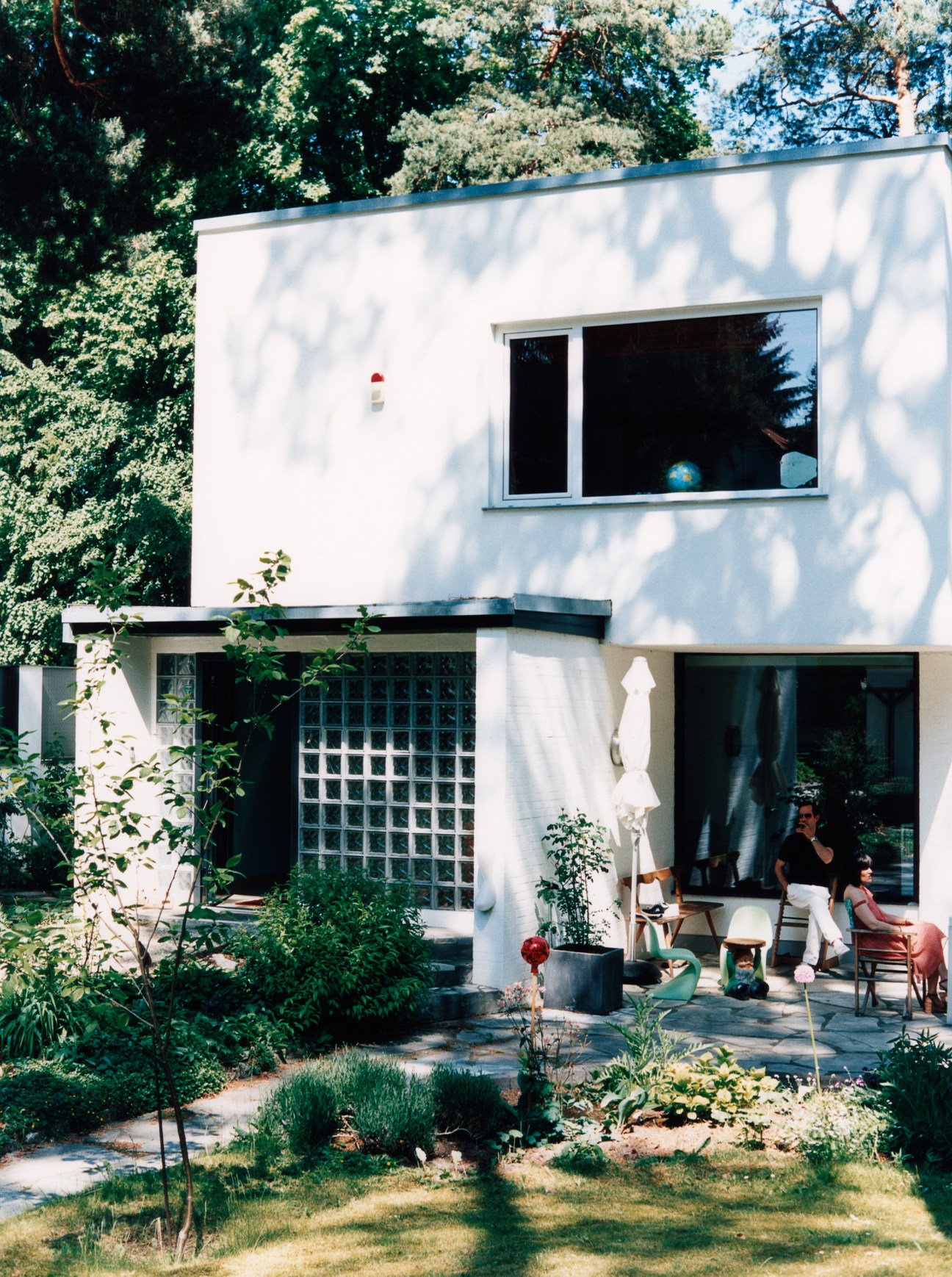 A modernist white house with flat roof and large windows, surrounded by lush green trees and garden.