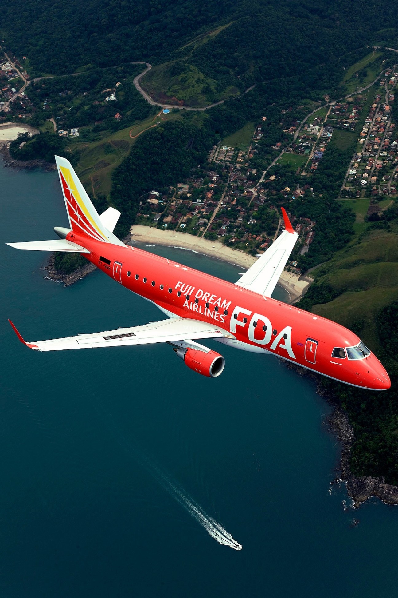 Red Fuji Dream Airlines aircraft flying over coastal landscape with turquoise water and forested terrain.