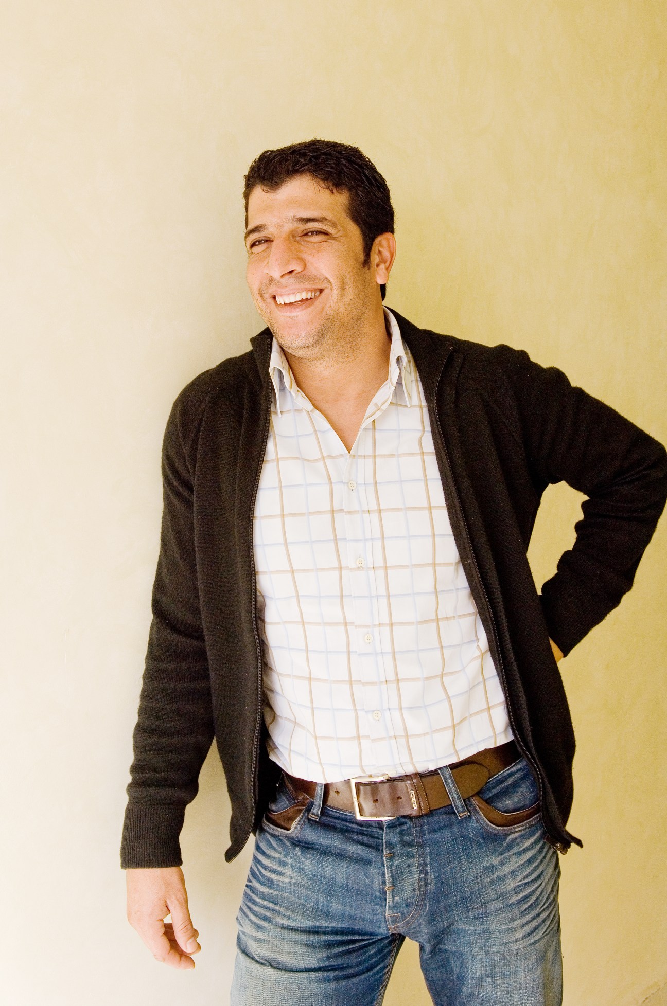 Portrait of Karim Toraif, a smiling man wearing a black cardigan and checkered shirt against a yellow background.
