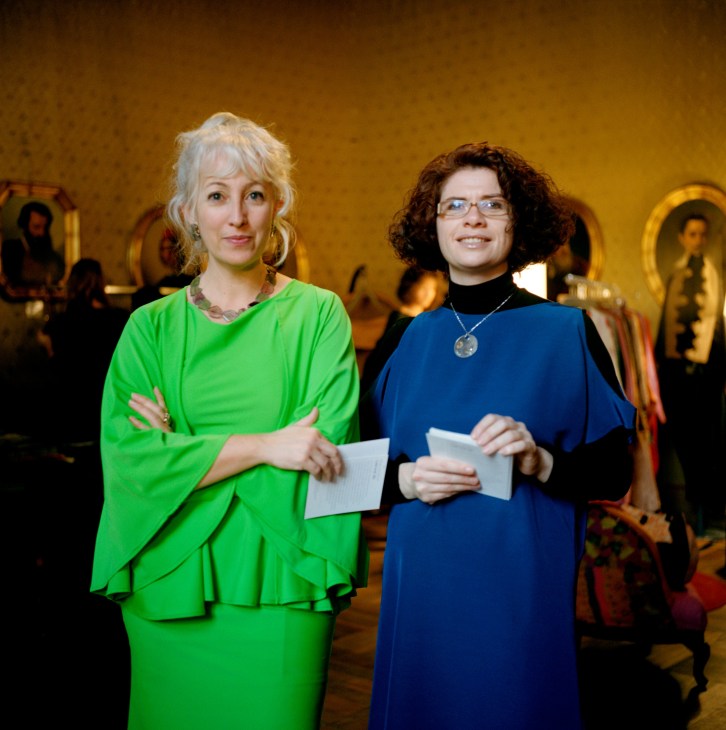 Two women standing in an ornate gallery with gold-framed portraits on yellow walls; one wears a bright green outfit and the other wears a dark blue dress.
