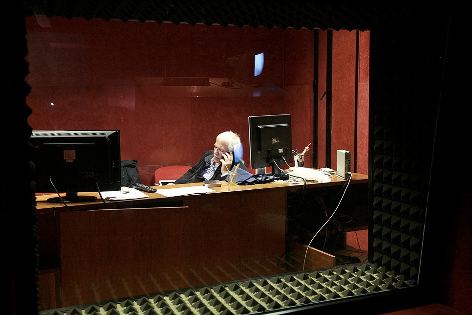 An older man sits at a desk in a red-walled recording studio, speaking on a telephone.