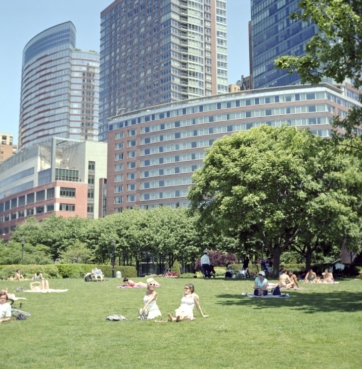 People relaxing on a grassy lawn with tall residential and office buildings in the background on a sunny day.