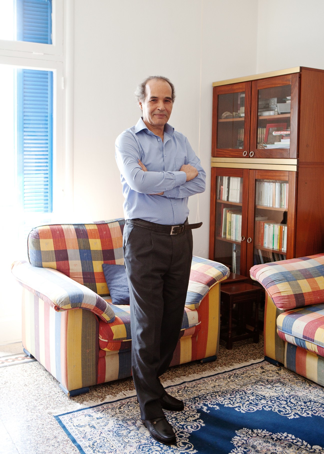 Mokhtar Yahyaoui stands with arms crossed in a bright room with bookshelves and colorful patterned furniture.