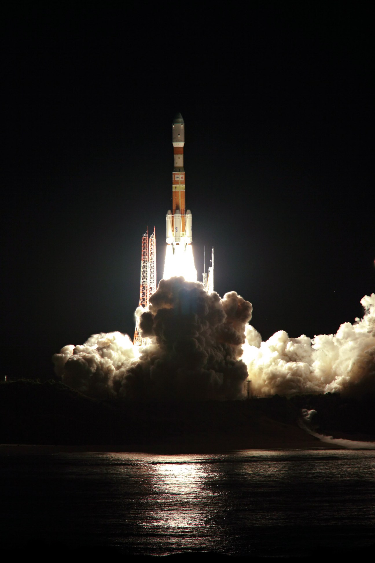 H-IIB rocket launching at night with bright flames and white smoke clouds billowing around the launch pad.
